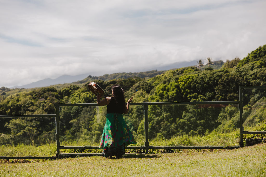 A woman in a colorful dress stands by a railing, holding a feather and facing a lush, green landscape with trees and distant mountains under a partly cloudy sky.