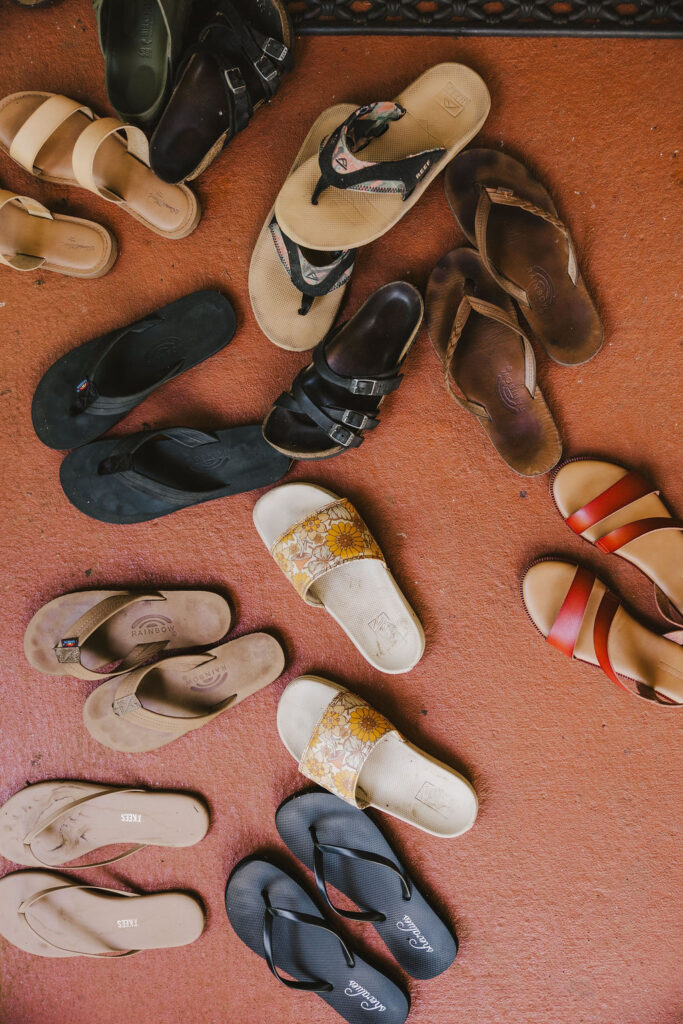 A variety of sandals and flip-flops, in different colors and styles, are scattered on a reddish-brown floor. The footwear appears to be for adults and is arranged in a semi-circle.