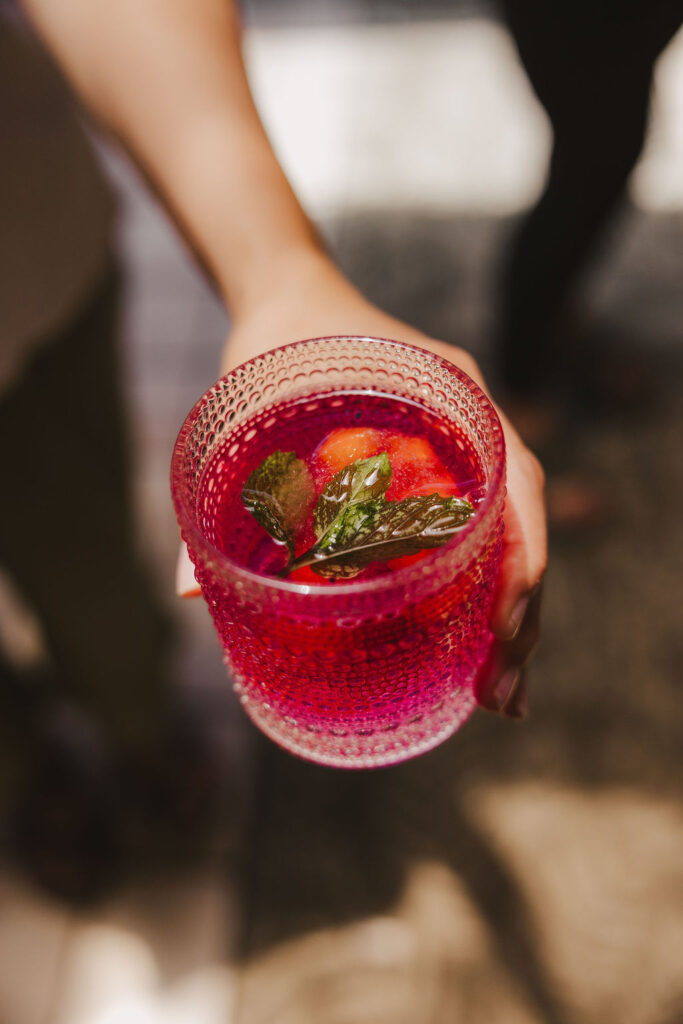 A hand holds a textured glass filled with a vibrant pink-red drink, garnished with mint leaves and a piece of fruit, possibly strawberry, with a blurred background.