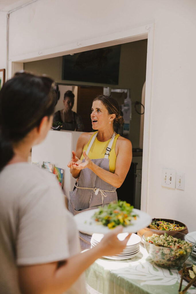Chef Angel Green, in a striped apron, gestures while speaking in a kitchen, standing behind a table with salads. Another person in the foreground holds a plate of food, attentively listening to her.