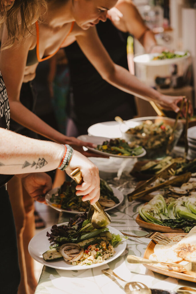 People serve themselves salad and other food from a buffet table filled with various fresh dishes. Sunlight shines on the table, and hands are seen holding plates and serving utensils. The atmosphere appears casual and social.