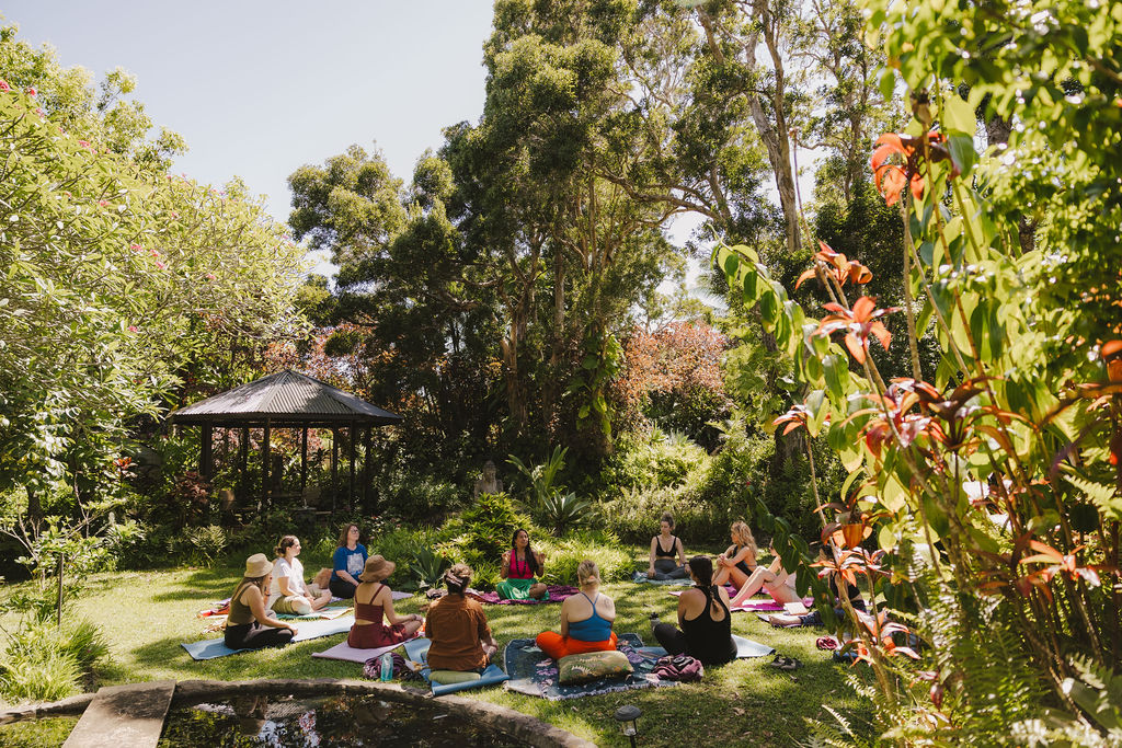 A group of people practice yoga or meditation on mats arranged in a circle on a grassy lawn, surrounded by lush greenery and tall trees, with a small gazebo in the background under a bright, sunny sky.