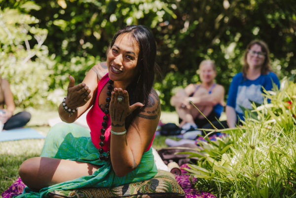 A woman with long hair and tattoos gestures and smiles while sitting on a mat outdoors, surrounded by greenery, with a small group of people sitting on the grass in the background.