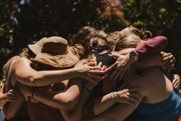 A group of people stand outdoors, embracing each other in a close, supportive group hug. Their faces are hidden and their arms are wrapped around one another, conveying a sense of unity and solidarity.