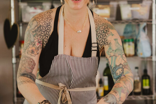 Chef Angel Green, a smiling person with tattoos and hoop earrings, wearing a striped apron, prepares sandwiches in a kitchen with shelves of ingredients in the background.