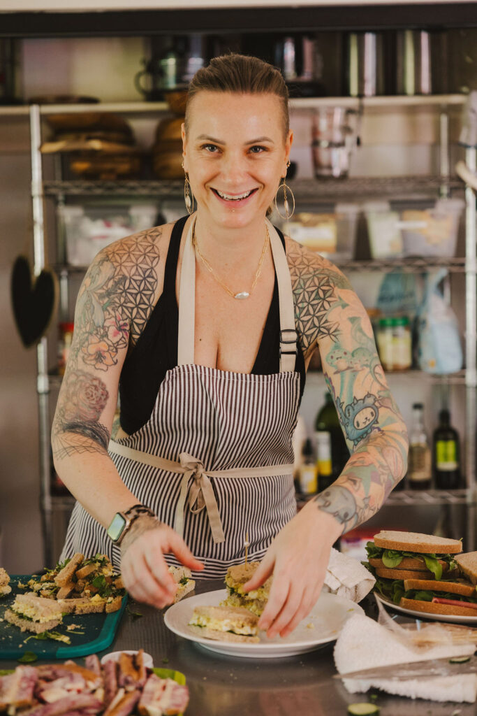 A smiling person with tattoos, wearing a striped apron, prepares sandwiches in a kitchen with shelves of ingredients and utensils in the background.