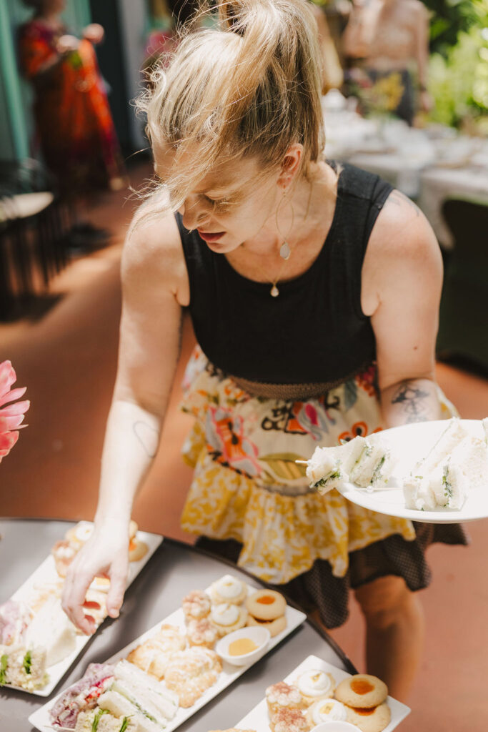 Chef Angel Green, a woman with blonde hair in a ponytail, wearing a black top and a colorful skirt, serves finger sandwiches and snacks onto a plate at an outdoor gathering.