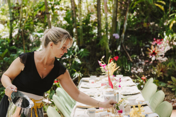 A woman in a black top and patterned skirt, part of the lotus chefs team, sets a long outdoor dining table with flowers, teacups, and plates under a glass pergola surrounded by lush greenery.