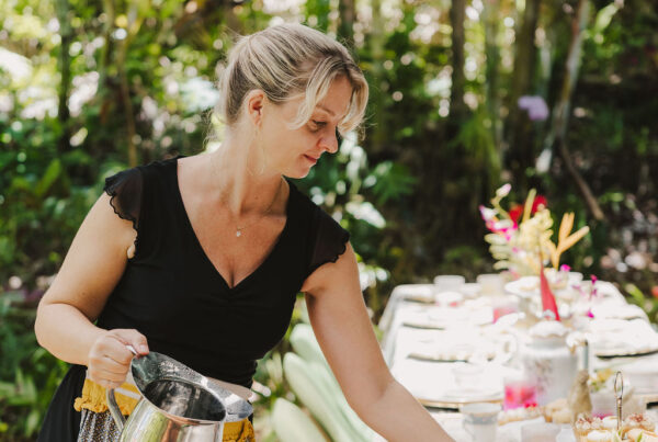 A woman with blonde hair, part of the lotus chefs team, pours water from a silver pitcher into a glass at an elegantly set outdoor table surrounded by greenery and flowers.