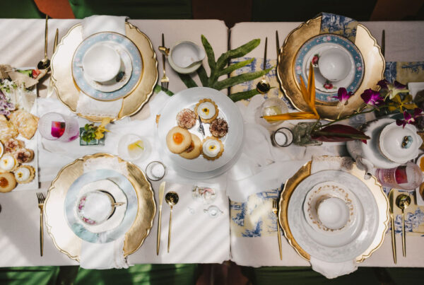 Elegant table setting for four with gold-trimmed plates, tea cups, and saucers. A tiered tray of assorted pastries is in the center, surrounded by flowers, drinks, and sunlight casting shadows over the scene.