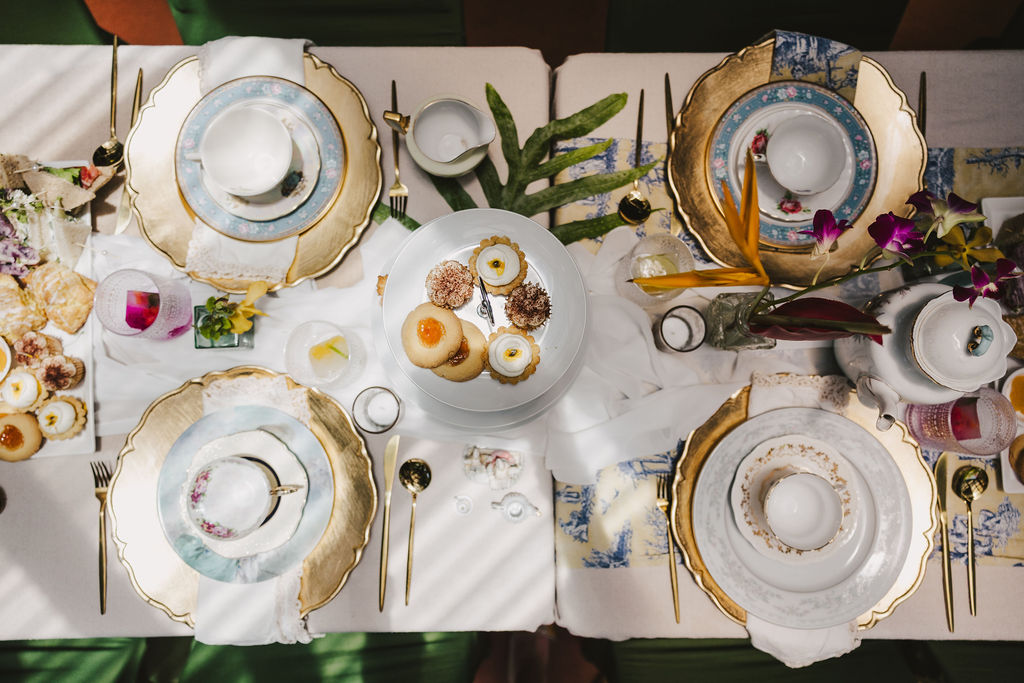 Elegant table setting for four with gold-trimmed plates, tea cups, and saucers. A tiered tray of assorted pastries is in the center, surrounded by flowers, drinks, and sunlight casting shadows over the scene.