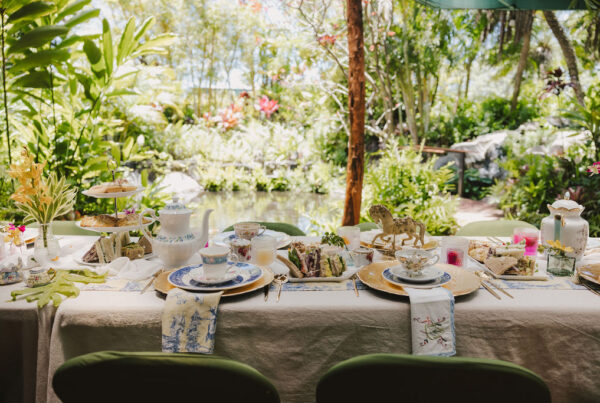 A beautifully set outdoor table for tea, decorated with elegant china, teapots, tiered trays of sandwiches and pastries, surrounded by lush greenery and a pond in the background.