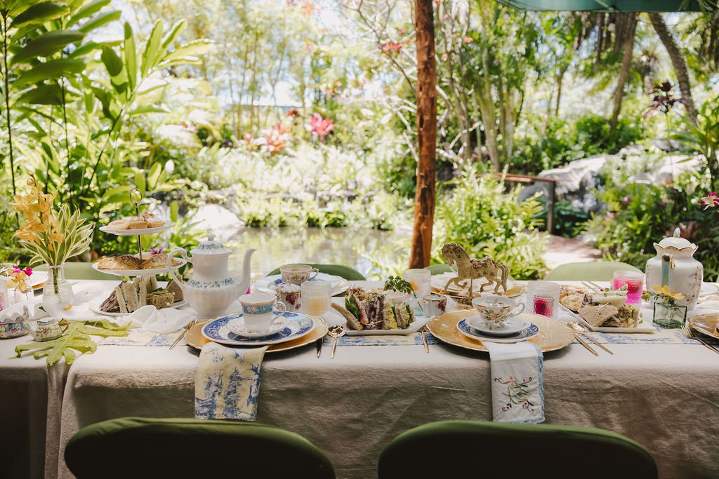 A beautifully set outdoor table for tea, decorated with elegant china, teapots, tiered trays of sandwiches and pastries, surrounded by lush greenery and a pond in the background.