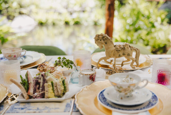 A close-up of an elegant outdoor table setting with vintage teacups, plates, finger sandwiches, and a decorative golden horse centerpiece, surrounded by lush greenery in the background.