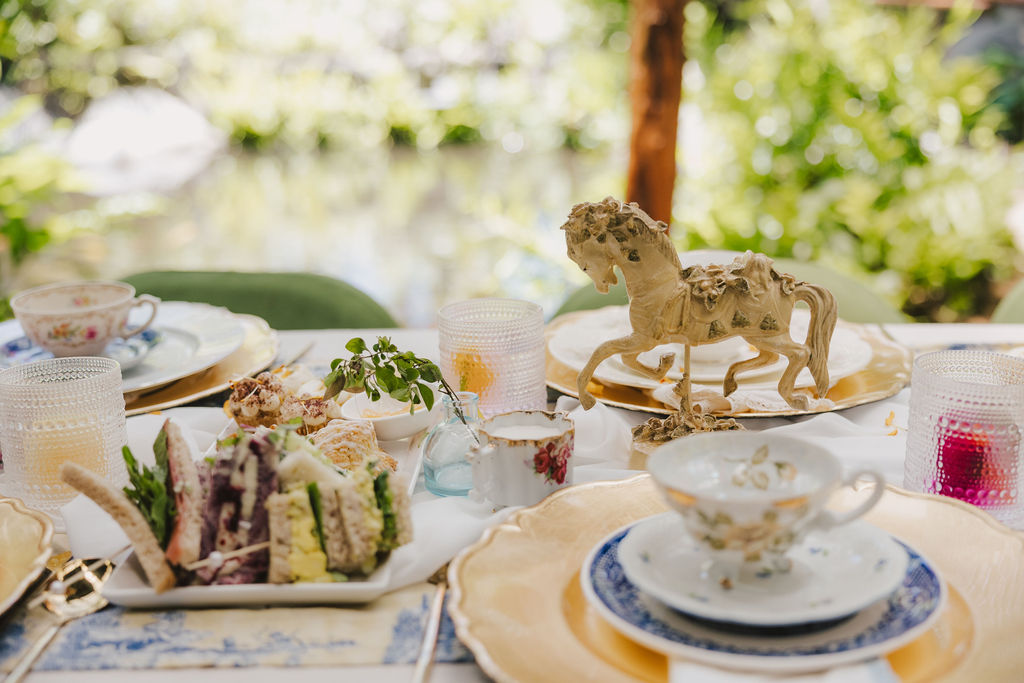 A close-up of an elegant outdoor table setting with vintage teacups, plates, finger sandwiches, and a decorative golden horse centerpiece, surrounded by lush greenery in the background.