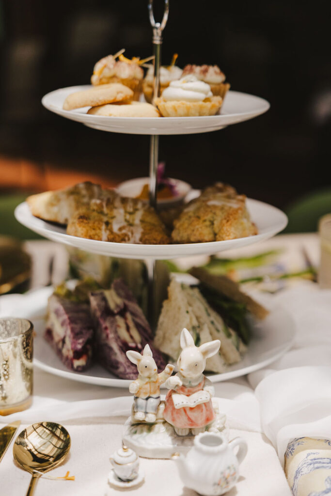 A three-tiered serving tray with assorted pastries, scones, and finger sandwiches on a table set for tea, with a small ceramic bunny figurine and miniature tea set in the foreground.