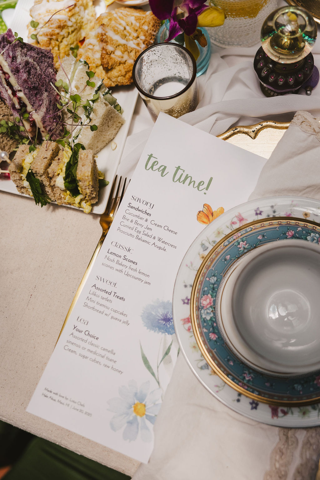 A tea party scene with elegant floral china, a teacup and saucer, cutlery, floral napkins, a menu labeled tea time!, and a plate of finger sandwiches and colorful scones on a cream tablecloth.