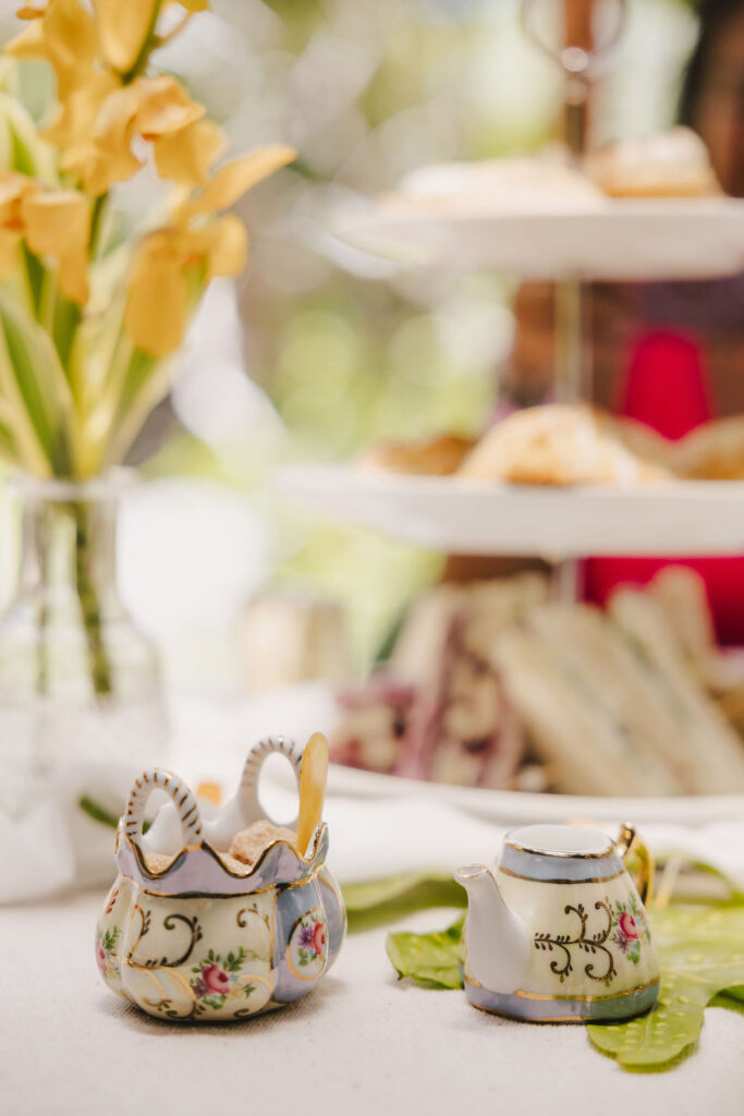A close-up of a decorative floral sugar bowl with spoons and a matching teapot on a table, with a vase of yellow flowers and a tiered tray of pastries in the background.