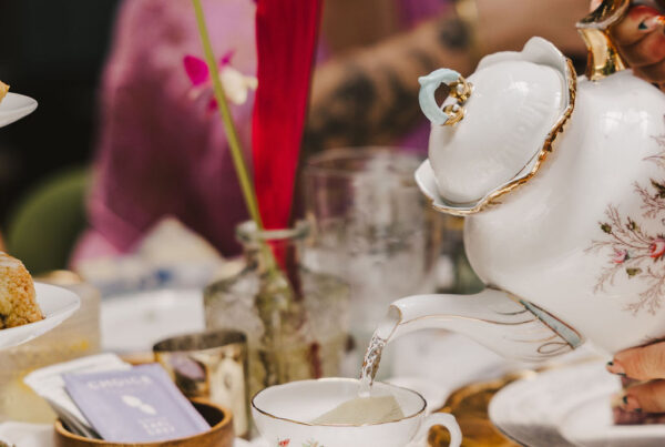 A close-up of a hand pouring tea from a floral teapot into a matching teacup at a decorated table, with colorful flowers and pastries, while people sit blurred in the background.