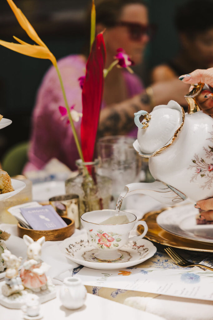 A close-up of a hand pouring tea from a floral teapot into a matching teacup at a decorated table, with colorful flowers and pastries, while people sit blurred in the background.