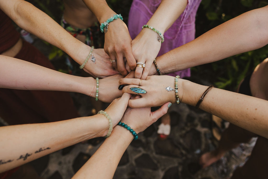 A group of people stand in a circle, each reaching into the center with one hand stacked atop another. Their arms show various bracelets, rings, and jewelry, with a stone path and greenery in the background.