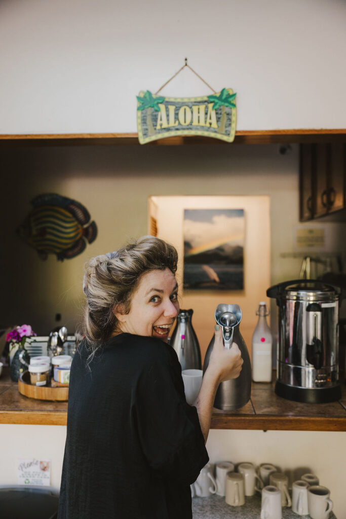 A smiling person with messy hair, wearing a black top, holds a metal whipped cream dispenser in a cozy kitchen decorated with mugs, flowers, and a sign reading ALOHA above the counter.