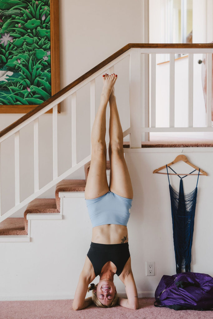 A woman practices a headstand against stairs indoors, wearing a black sports bra and light blue shorts. A blue dress hangs on the railing, and a purple bag sits on the floor nearby. A painting decorates the wall.