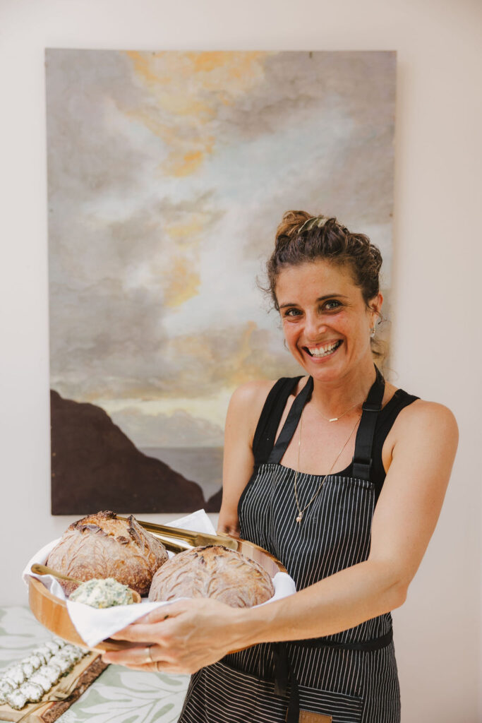 Chef Angel Green, smiling in a striped apron, holds a tray with rustic bread and a bowl of spread, standing indoors in front of a large painting of a cloudy sky over hills.