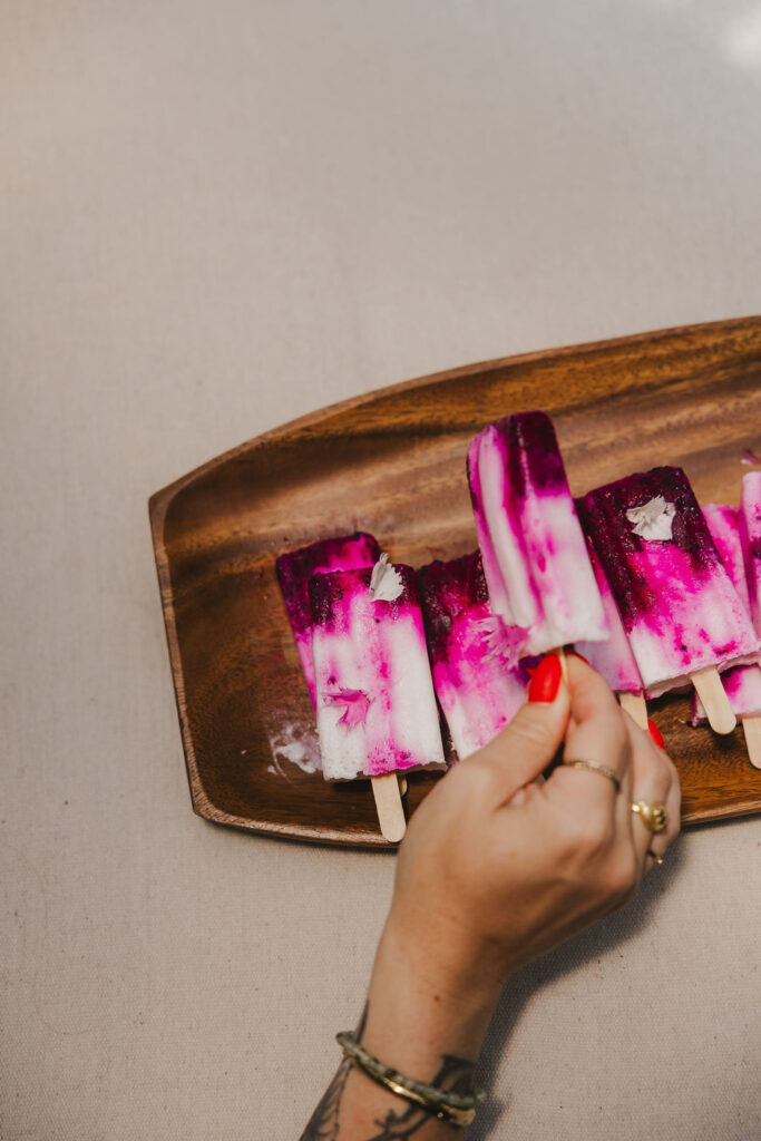 A hand with red nail polish holds a vibrant pink and white popsicle from a wooden tray containing several similar popsicles, on a light beige surface.