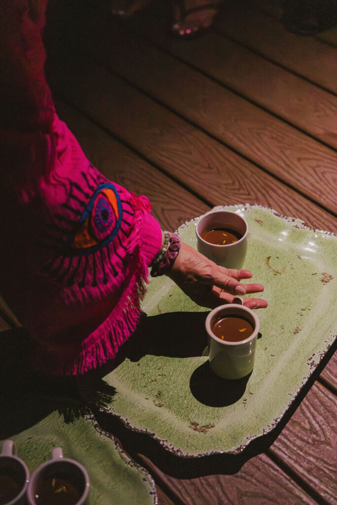 A person wearing a bright pink garment with a blue and red eye design reaches for a white mug of tea on a green tray, with two other mugs also on the tray, set on a wooden floor.