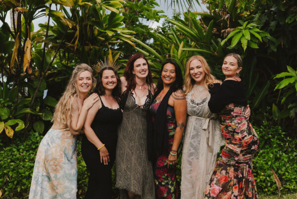 Six women stand closely together, smiling and posing in front of lush green tropical plants. They are dressed in colorful and elegant dresses, creating a celebratory and joyful atmosphere.