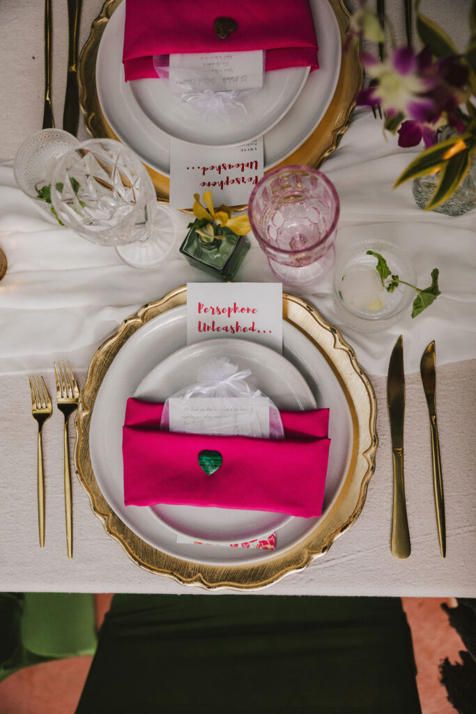 A formal table setting with gold-rimmed plates, gold cutlery, crystal glasses, and a hot pink napkin on each plate. A place card reading Persephone Unleashed sits atop each napkin.