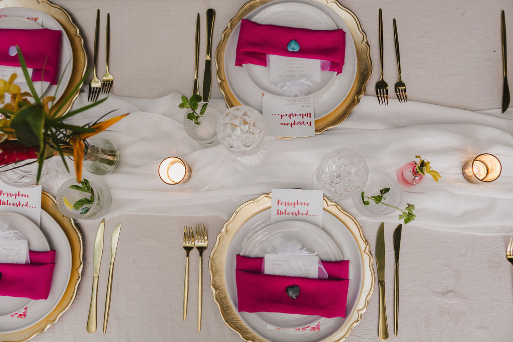 Elegant table setting with gold-rimmed plates, bright pink napkins, handwritten place cards, gold cutlery, crystal glasses, small floral arrangements, and candles on a white tablecloth with a sheer runner.
