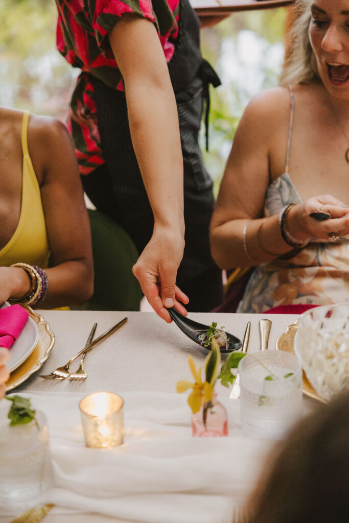 A server places a small dish in front of a smiling woman at a decorated dining table with candles, flowers, and glassware. Two women in colorful dresses sit at the table.