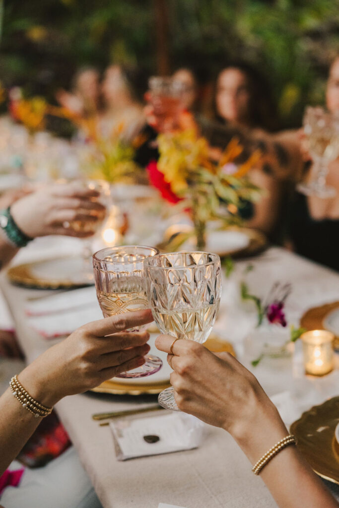 A group of people at a decorated table raise crystal glasses in a toast, surrounded by flowers, candles, and elegant place settings at an outdoor gathering.