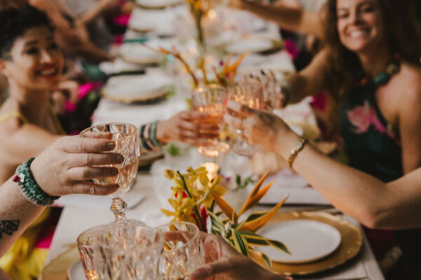 A group of people sitting around a long, decorated table outdoors, smiling and raising crystal glasses for a toast. The table is set with plates, gold chargers, flowers, and candles.