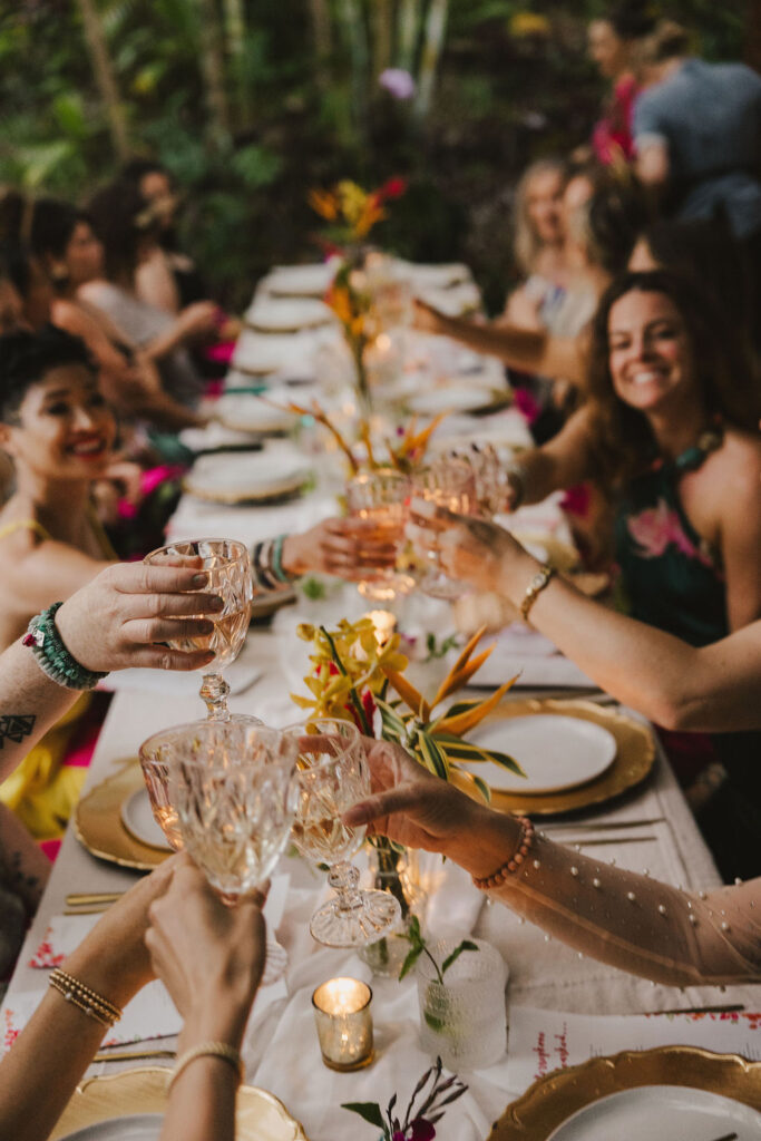 A group of people sitting around a long, decorated table outdoors, smiling and raising crystal glasses for a toast. The table is set with plates, gold chargers, flowers, and candles.