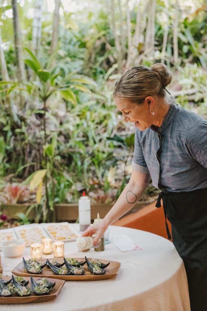 A woman wearing a blue shirt and black apron smiles while preparing plates of food on a round table outdoors, surrounded by lush greenery and lit candles.