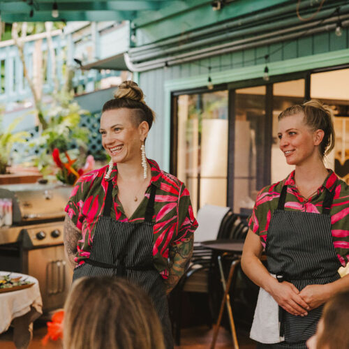 Two women, including Tara Klitchman, wearing matching pink and black shirts with aprons, stand smiling in an outdoor kitchen area. A grill, table of food, and bottles are visible in the background as people sit in the foreground.