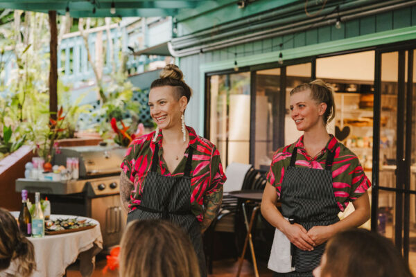 Two women in matching pink and black shirts with aprons smile while standing in an outdoor kitchen area, with food and drinks on a table beside them and greenery in the background.