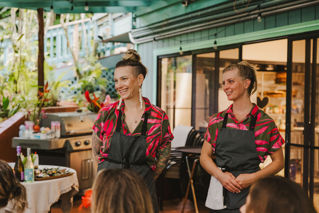 Two women in matching pink and black shirts with aprons smile while standing in an outdoor kitchen area, with food and drinks on a table beside them and greenery in the background.