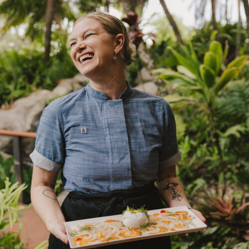 A smiling chef in a blue shirt and black apron stands outdoors surrounded by lush greenery, holding a rectangular plate with a beautifully arranged dish.