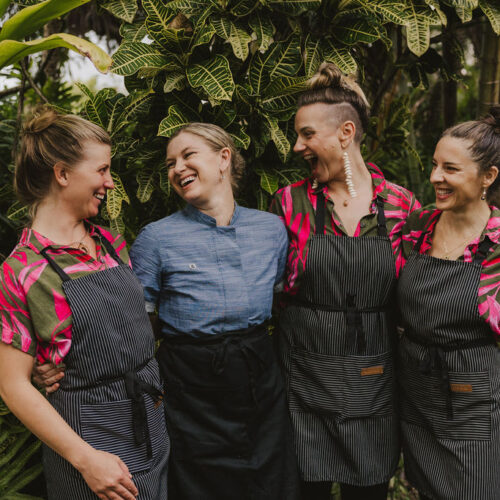 Four women, including Tara Klitchman, stand together outdoors, smiling and laughing. Three wear matching pink floral shirts with striped aprons, while one sports a blue chef coat and black apron amid lush green foliage.