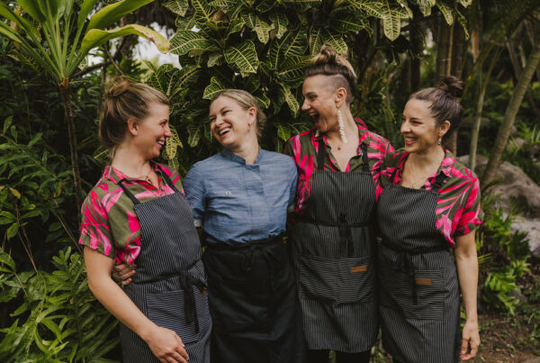 Four women stand smiling together in a lush, tropical garden. Three wear pink floral shirts with striped aprons, while the fourth wears a blue chef’s jacket and black apron. They appear happy and relaxed among greenery.
