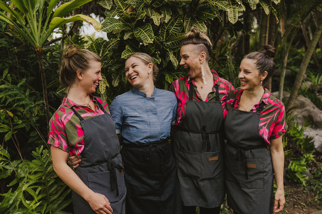 Four women stand smiling together in a lush, tropical garden. Three wear pink floral shirts with striped aprons, while the fourth wears a blue chef’s jacket and black apron. They appear happy and relaxed among greenery.