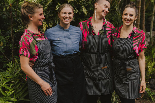 Four women stand together smiling in a lush garden. Three wear pink and black print shirts with striped aprons, while the fourth wears a blue shirt and a black apron. They appear to be enjoying each others company.