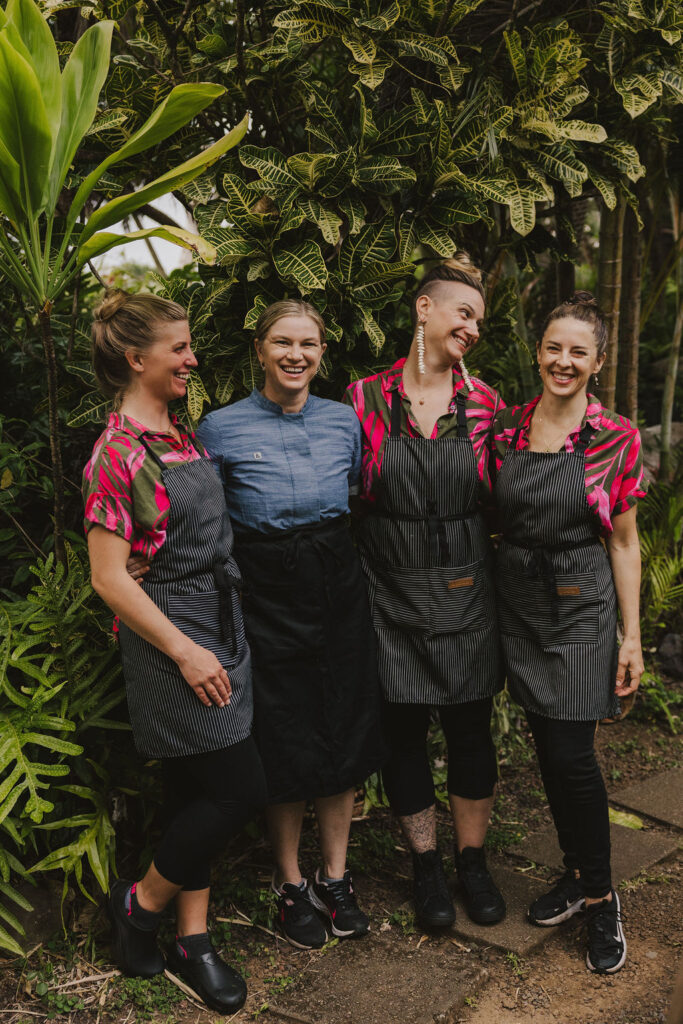 Four women stand together smiling in a lush garden. Three wear pink and black print shirts with striped aprons, while the fourth wears a blue shirt and a black apron. They appear to be enjoying each others company.