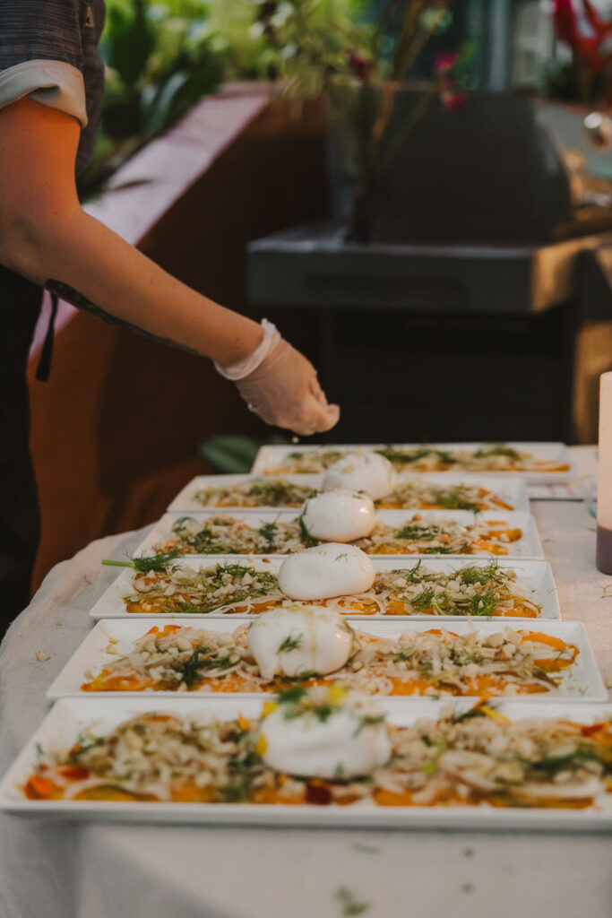 A person wearing a glove is preparing and garnishing multiple plates of burrata cheese with vegetables and herbs on a table. The plates are neatly arranged in rows, ready to be served.