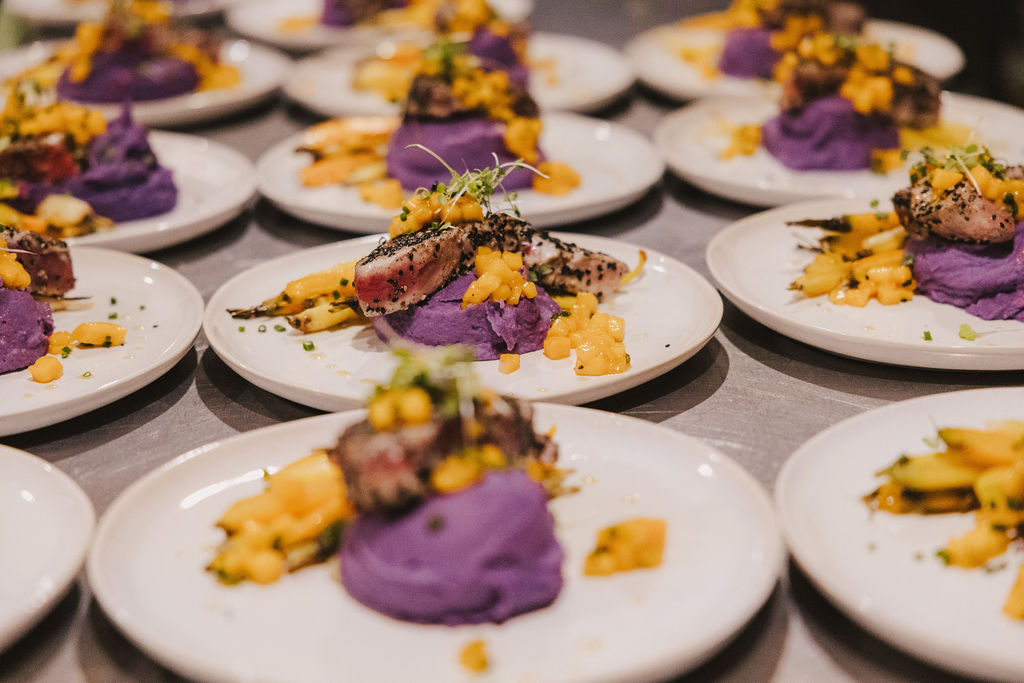 Multiple white plates each with a serving of purple mashed potatoes, seared fish topped with herbs, and a yellow vegetable or fruit relish, arranged in rows on a table.