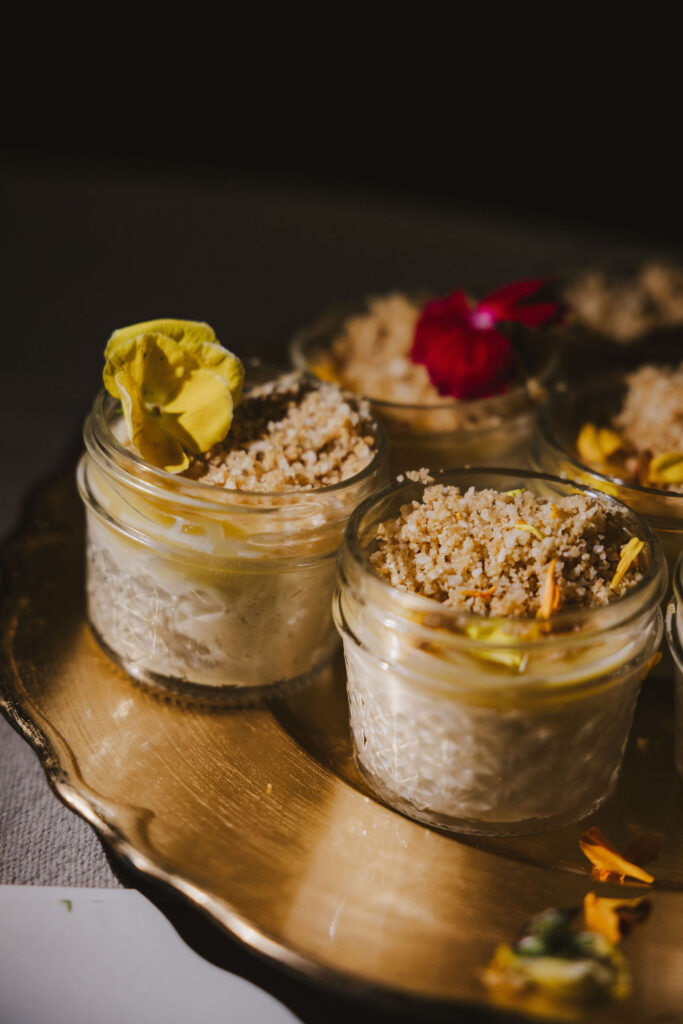 Small glass jars filled with creamy dessert, topped with crumbled topping and garnished with colorful edible flowers, arranged on a gold tray.